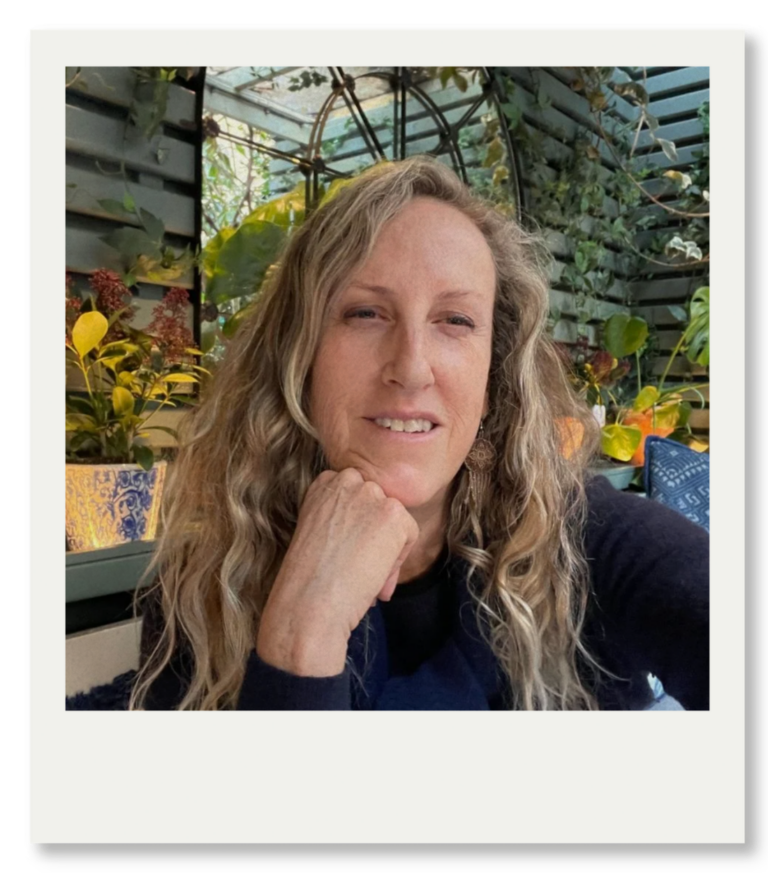 Sarah M Rubin seated in a plant‑filled outdoor space, resting her chin on her hand.