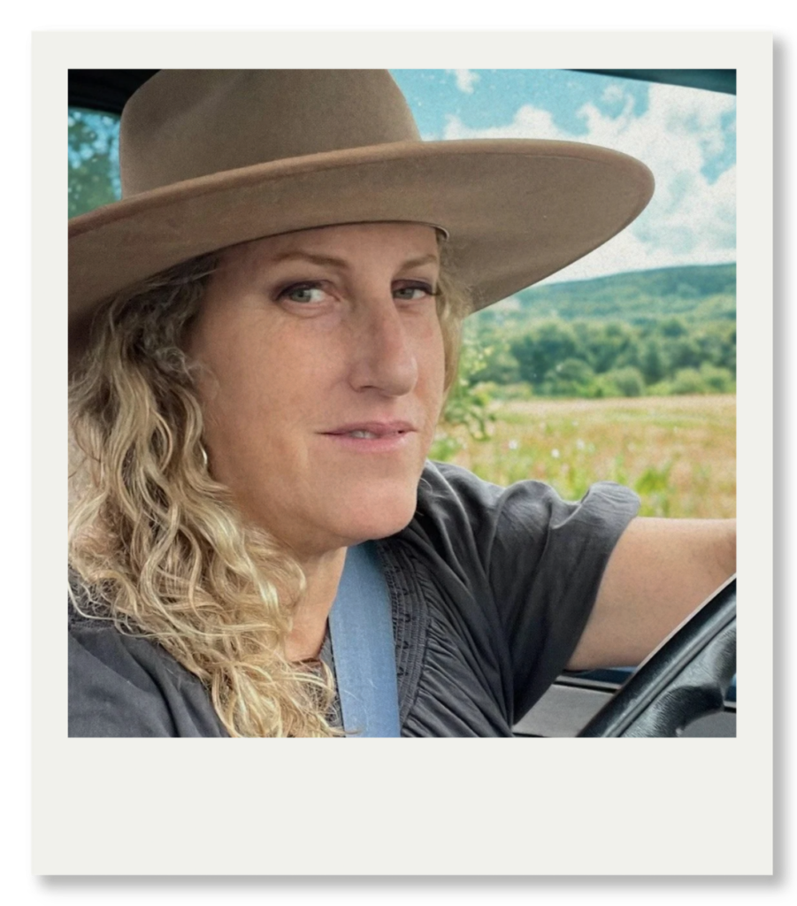 Sarah M Rubin driving, wearing a wide‑brimmed hat, with fields and hills visible through the window.